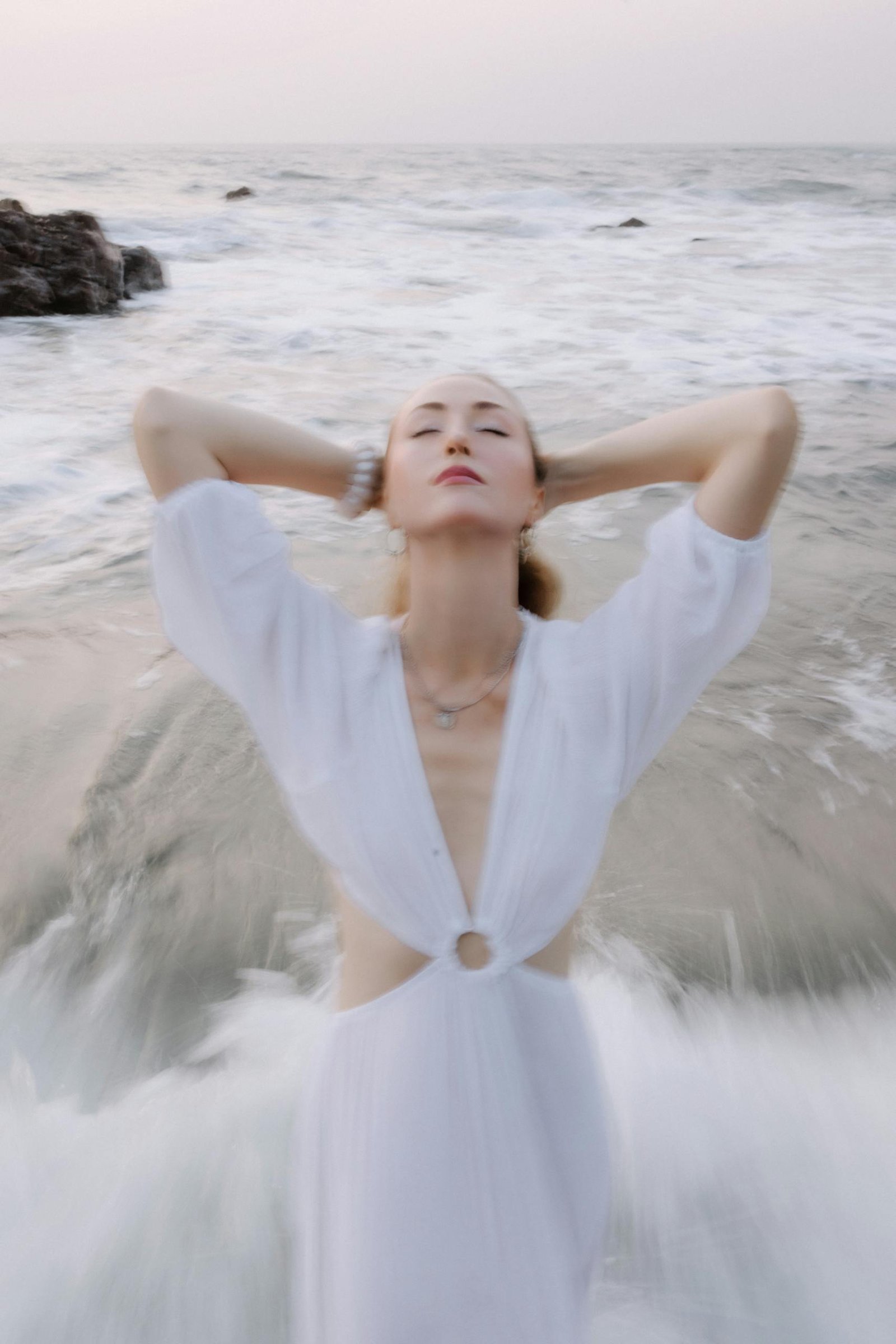 A healthy woman in a white dress enjoys the ocean breeze, standing in gentle waves.
