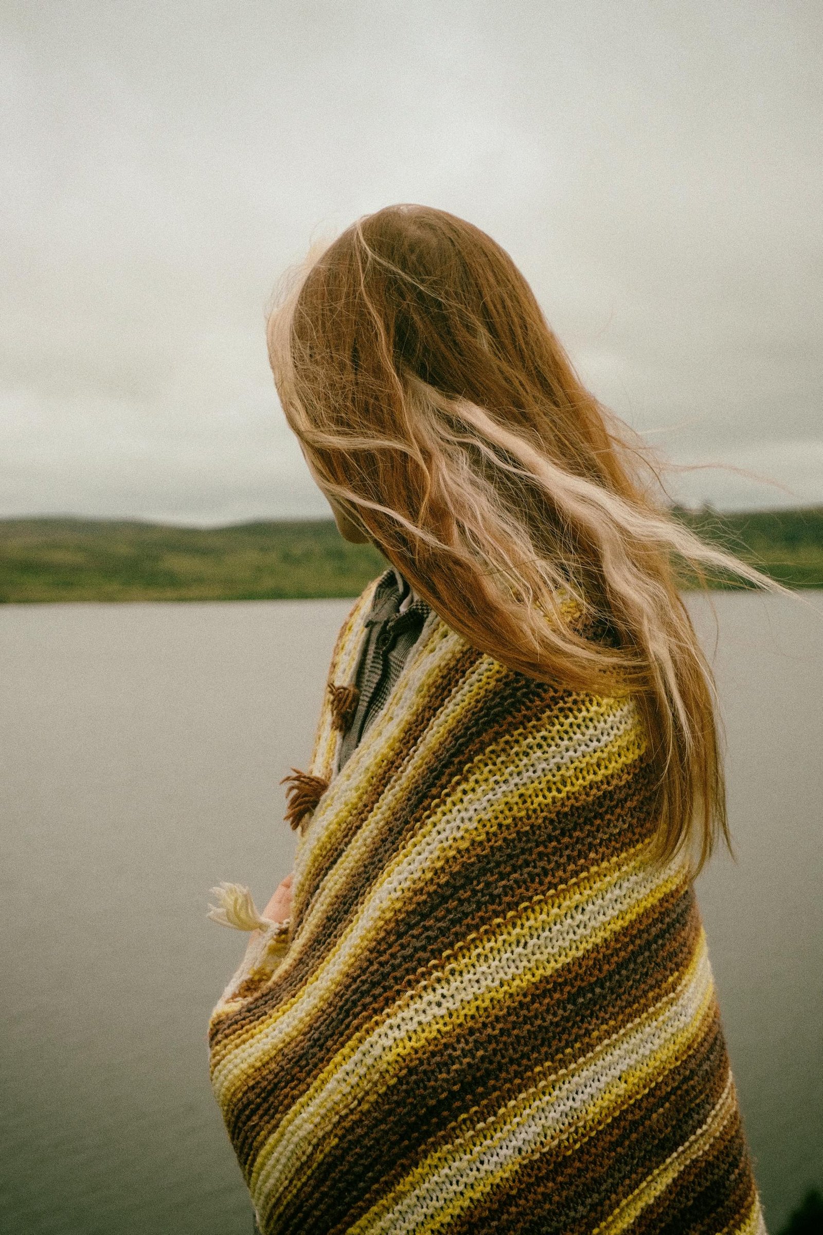 A woman wrapped in a shawl gazes at the calm lake under a cloudy sky.